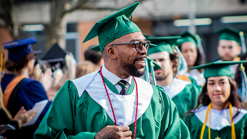 A group of graduates in green caps and gowns walking during a graduation ceremony, with one man in the foreground smiling and adjusting his cap.