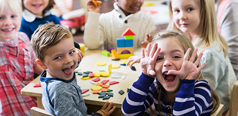 A group of very young children smiling and laughing together at a table with colorful, wooden building blocks