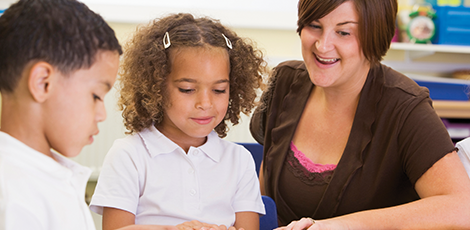 Two elementary schoolers being assisted by a teacher while reading a book