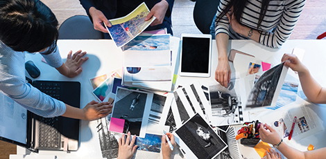 People sitting around a table cluttered with printed out pictures, and laptops