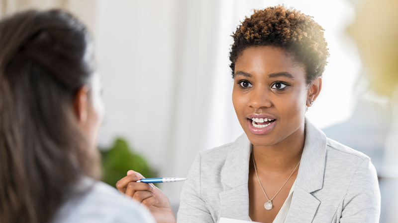 A woman in a light gray blazer speaks while holding a pen, seated across from another woman in a bright office with a blurred plant in the background.